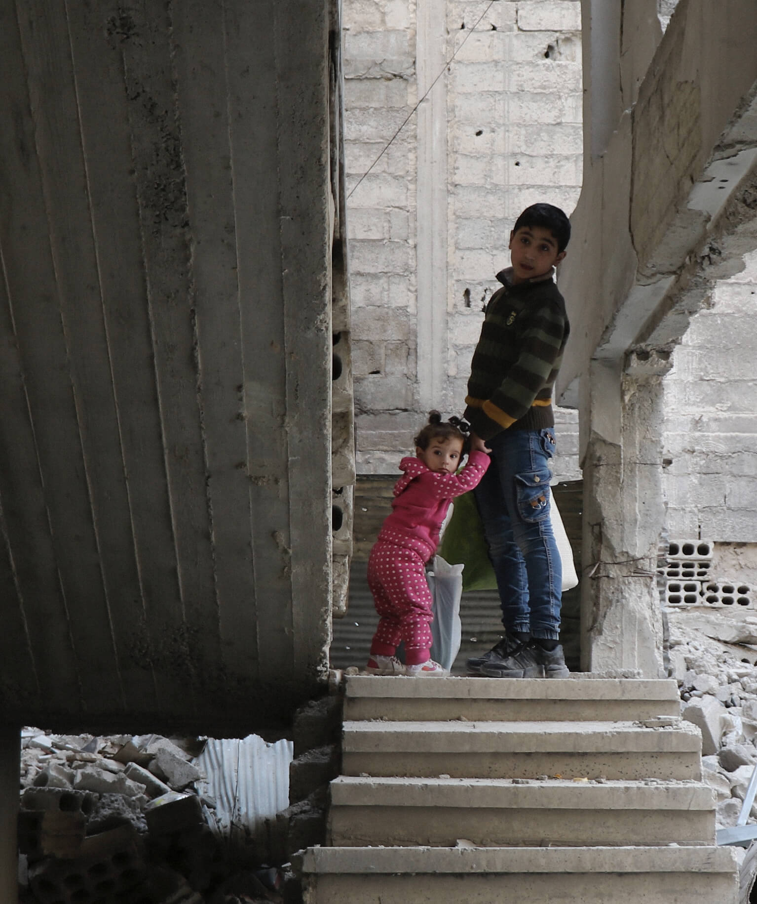 two children holding hands walking up stairs surrounded by rubble
